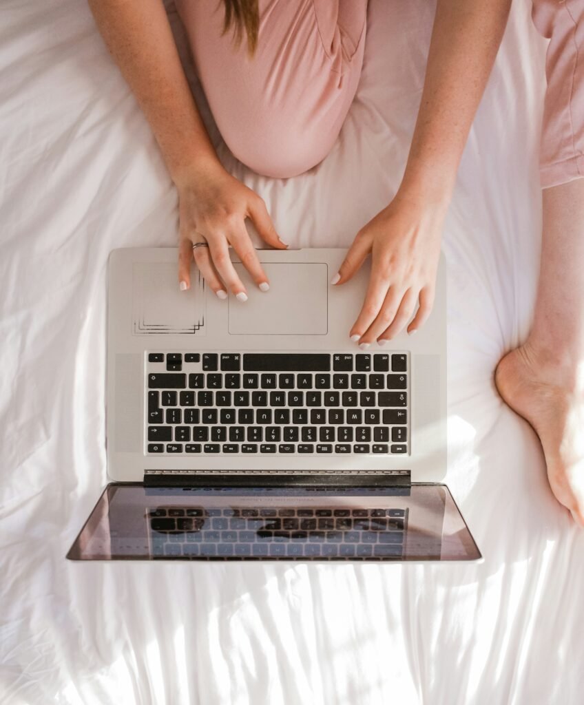 Woman in pink pyjamas typing on a laptop while sitting on a white bed